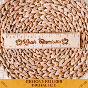 May include: A wooden ruler with a smiley face and the names "Leah Charlotte" engraved on it. The ruler is 8 inches long and has markings for each inch. The ruler is on a woven surface.