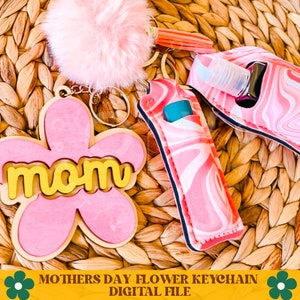 May include: Pink and yellow flower keychain with the word "mom" on it. The keychain is attached to a pink and white patterned lip balm holder and a pink fuzzy pom-pom.