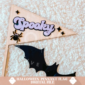 K&ouml;nnte beinhalten: Eine Holz-Wimpelflagge mit dem Text "Spooky" in Lila und einer schwarzen Spinne mit wei&szlig;en Augen. Die Flagge ist an einem schwarzen Fledermaus-Ausschnitt befestigt.