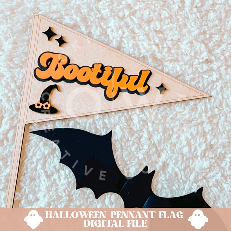 K&ouml;nnte beinhalten: Eine Holz-Wimpelflagge mit dem Wort "Bootiful" in orangefarbenen Buchstaben mit schwarzen Umrandungen. Unter der Flagge befindet sich eine schwarze Fledermaus-Silhouette. Die Flagge liegt auf einer wei&szlig;en, flauschigen Oberfl&auml;che.