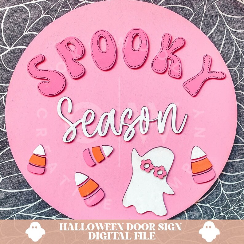 Peut inclure: Une pancarte ronde en bois rose avec les mots "Spooky Season" en lettres blanches et roses. La pancarte pr&eacute;sente &eacute;galement un fant&ocirc;me avec des lunettes de soleil roses et du ma&iuml;s sucr&eacute;.