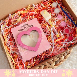 May include: A pink wooden card with a heart-shaped cutout and the text "Happy Mother's Day" is in a box with colorful shredded paper. The box also contains paint, a paintbrush, and a candle.