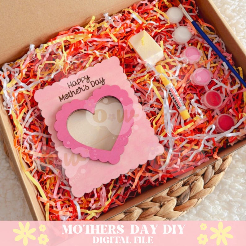 May include: A pink wooden card with a heart-shaped cutout and the text "Happy Mother's Day" is in a box with colorful shredded paper. The box also contains paint, a paintbrush, and a candle.