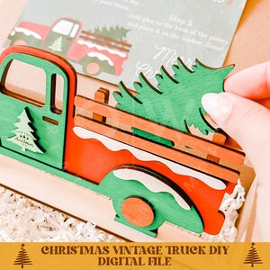 May include: A red vintage truck with a green Christmas tree in the bed. The truck is made of wood and has a white snow design on the sides. The truck is a DIY project and comes with instructions. The instructions say "Add glue to the back of the pieces and place it on the bucker. Done!"