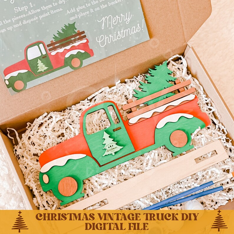 May include: A Christmas vintage truck DIY kit with a red and green wooden truck carrying a Christmas tree. The kit includes paint brushes and a wooden base. The box is open and the truck is sitting on a bed of shredded paper. The box lid has a Merry Christmas message.