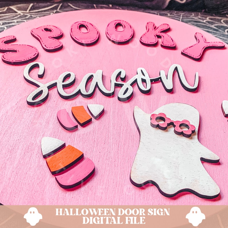 Peut inclure: Une pancarte de porte ronde en bois rose avec les mots "Spooky Season" en lettres blanches. La pancarte pr&eacute;sente &eacute;galement un fant&ocirc;me blanc avec des lunettes roses et deux d&eacute;corations de ma&iuml;s sucr&eacute; roses et orange.