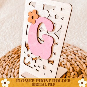 Op de afbeelding: Houten telefoonhouder met een bloemenontwerp en de letter "G" in roze. De letter is versierd met een oranje bloem. De houder heeft een uitgesneden bloemenpatroon en een standaard. De tekst "FLOWER PHONE HOLDER DIGITAL FILE" staat onderaan.