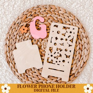 Op de afbeelding: Een houten telefoonhouder met een bloemenontwerp, een aparte houten basis en een roze vilt letter "G" zijn gerangschikt op een geweven mat. Een oranje bloem en de tekst "FLOWER PHONE HOLDER DIGITAL FILE" zijn ook zichtbaar.