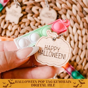 Op de afbeelding: Een houten sleutelhanger in de vorm van een pompoen met de tekst "Happy Halloween!" erop. De sleutelhanger heeft een groene en roze pop-it.