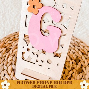 Op de afbeelding: Houten telefoonhouder met een lasergesneden bloemenontwerp. Voorzien van een roze letter "G" en een oranje bloemaccent. De basis heeft een sleuf voor het vasthouden van een telefoon. De tekst "FLOWER PHONE HOLDER DIGITAL FILE" staat onderaan.