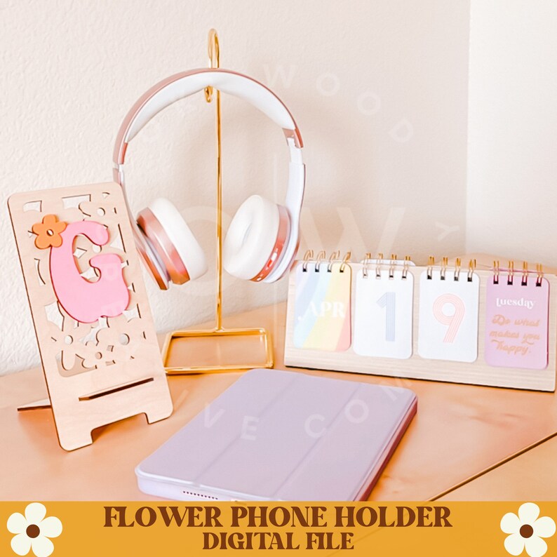 Op de afbeelding: Een houten telefoonhouder met een roze letter "G" en bloemdesign, een gouden hoofdtelefoonstandaard met witte en roze hoofdtelefoons, een houten kalender en een lila tablethoes. De kalender toont "19 april" en "dinsdag".