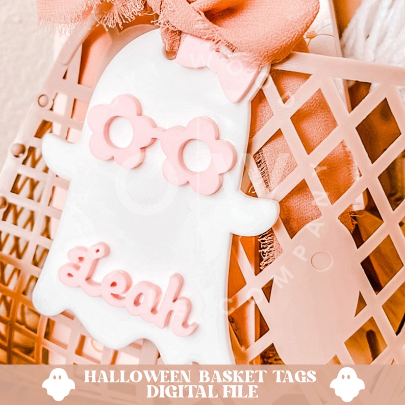 Pode incluir: Uma etiqueta de cesta de Halloween em forma de fantasma branco com &oacute;culos rosa e o nome "Leah" escrito em rosa. A etiqueta est&aacute; em uma cesta de arame rosa.