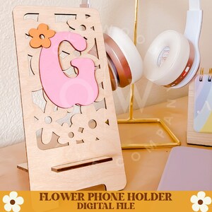 Op de afbeelding: Een houten telefoonhouder met een lasergesneden bloemenontwerp. Een roze letter "G" met een oranje bloemaccent is aan de voorkant bevestigd. De tekst "FLOWER PHONE HOLDER DIGITAL FILE" staat onderaan. Koptelefoon op de achtergrond.