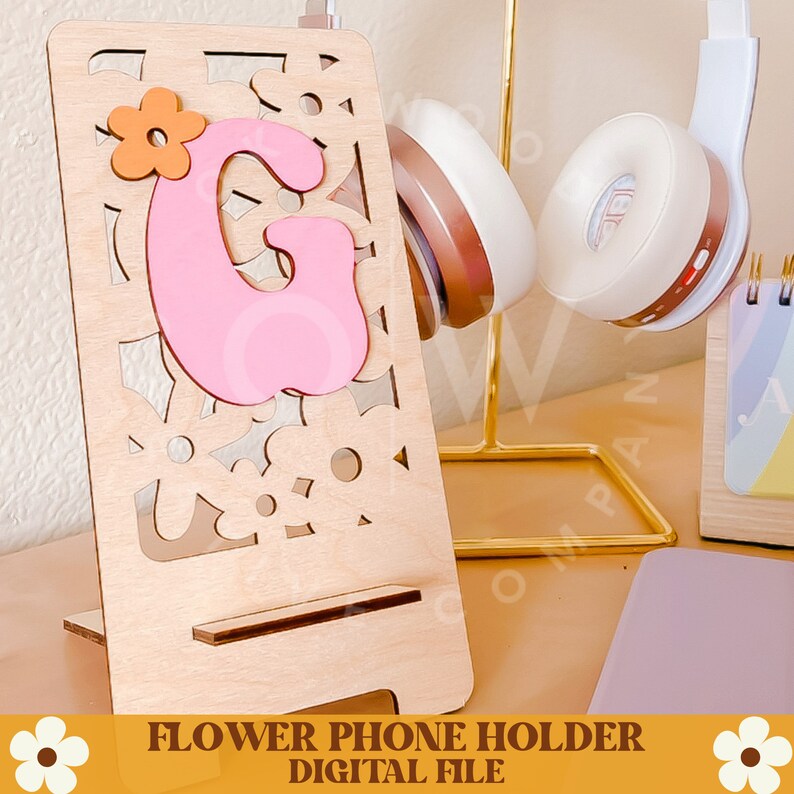 Op de afbeelding: Een houten telefoonhouder met een lasergesneden bloemenontwerp. Een roze letter "G" met een oranje bloemaccent is aan de voorkant bevestigd. De tekst "FLOWER PHONE HOLDER DIGITAL FILE" staat onderaan. Koptelefoon op de achtergrond.