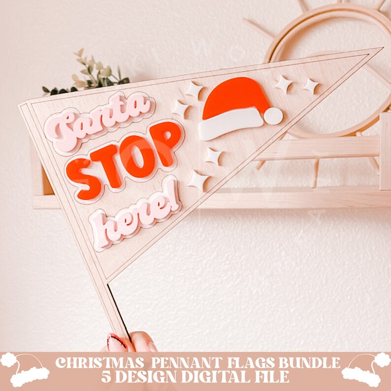 May include: A wooden triangle pennant flag with the words "Santa Stop Here!" in pink and red letters. A red Santa hat with white trim is on the flag. The flag is on a wooden shelf.