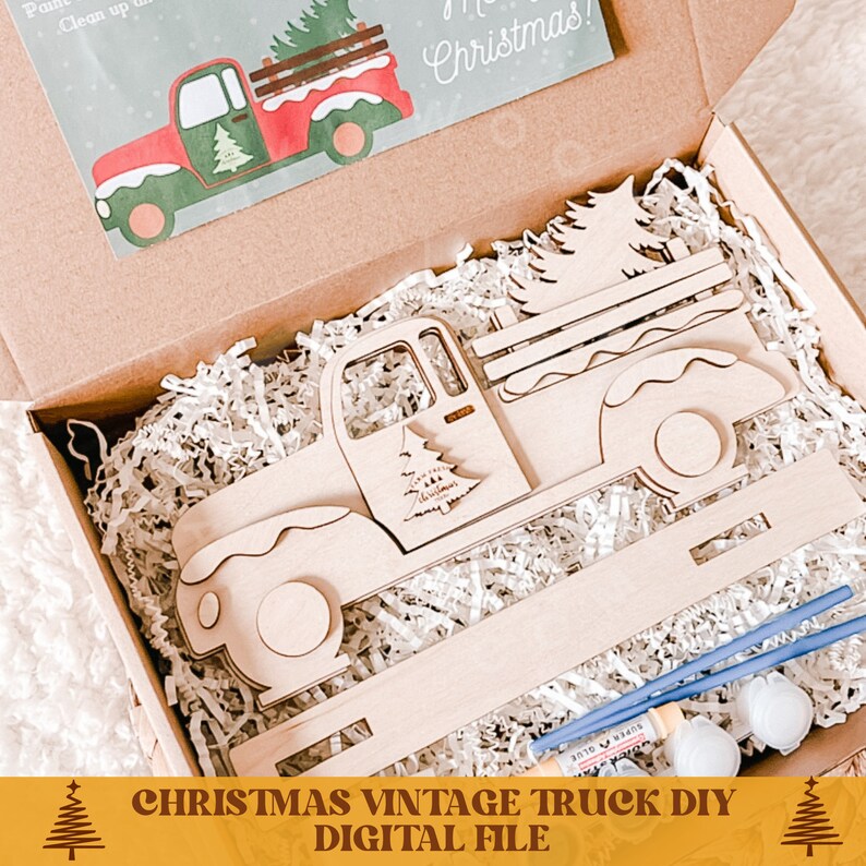 May include: A wooden Christmas truck cutout with a Christmas tree in the bed of the truck. The truck is in a brown box with white shredded paper. The box also contains paint and paintbrushes. The text "Merry Christmas!" is on a card in the box.