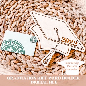 May include: A wooden graduation cap cutout with the year 2022 engraved on it. The cap is designed to hold a gift card. The image includes a Starbucks gift card as an example.  The text "Marketing Pictures Included" is also visible in the image.