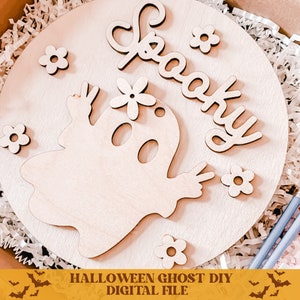 May include: A wooden round sign with a spooky ghost and the word "Spooky" in cursive. The ghost has a flower on its head and is holding up two fingers in a peace sign. There are small flowers around the ghost and the word "Spooky".