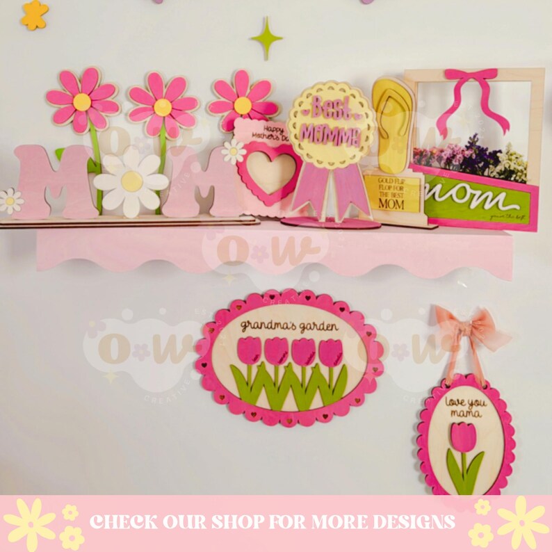 May include: A pink shelf with various wooden decorations for Mother's Day. The decorations include a sign that says "Mom", flowers, a heart, a ribbon with the text "Best Momma", a sign that says "Gold Star for the Best Mom", and a picture frame with the word "Mom" on it.