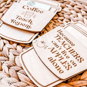 May include: Two wooden coffee cup-shaped signs with teacher-themed sayings. The first sign says "Coffee Teach Repeat" with a coffee cup icon. The second sign says "Because Teachers can't live on Apples alone" with an apple icon.
