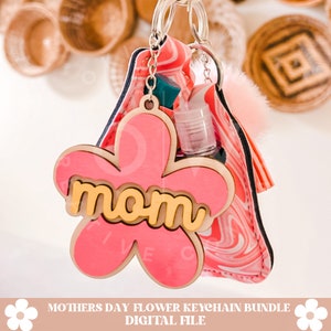Puede incluir: Llavero de madera con forma de flor rosa con la palabra "mom" en letras amarillas. El llavero est&aacute; unido a una bolsa de tela de color rosa y blanco con un bolsillo de pl&aacute;stico transparente para desinfectante de manos.