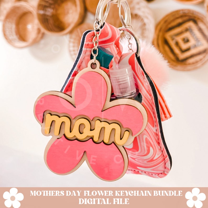 Puede incluir: Llavero de madera con forma de flor rosa con la palabra "mom" en letras amarillas. El llavero est&aacute; unido a una bolsa de tela de color rosa y blanco con un bolsillo de pl&aacute;stico transparente para desinfectante de manos.