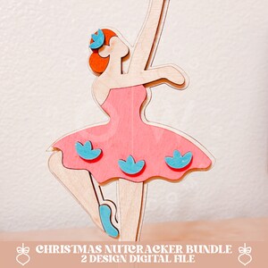 Pu&ograve; includere: Una statuetta di legno di un'omino di pan di zenzero a forma di ballerina con un vestito rosa con fiori blu. La ballerina ha i capelli rossi e una corona blu.