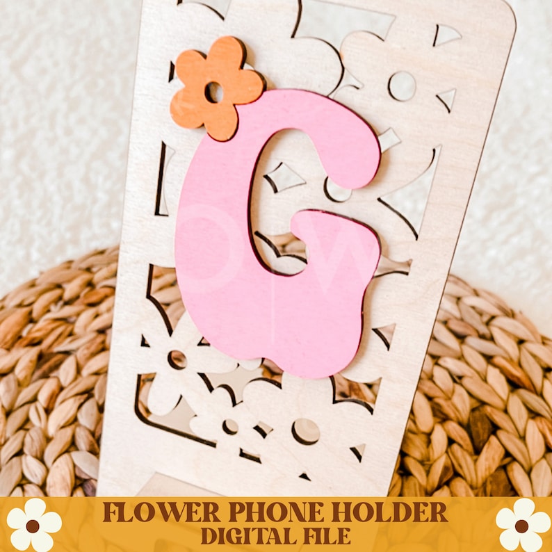 Op de afbeelding: Een houten telefoonhouder met een roze letter "G" en een oranje bloemaccent. De houder heeft een bloemenuitsparing en de tekst "FLOWER PHONE HOLDER DIGITAL FILE" onderaan. De houder staat tegen een geweven achtergrond.