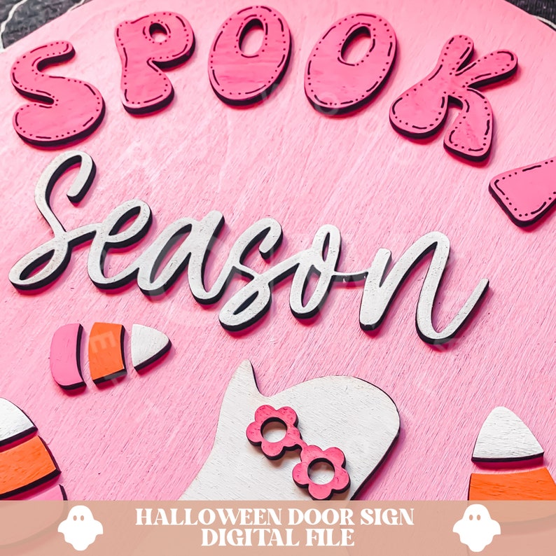Peut inclure: Une pancarte de porte en bois rose avec le texte "Spook Season" en lettres blanches et roses. La pancarte pr&eacute;sente &eacute;galement un fant&ocirc;me blanc avec des lunettes en forme de fleurs roses et deux d&eacute;corations de ma&iuml;s sucr&eacute;.