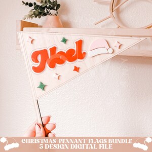 May include: A wooden triangle pennant flag with the word "Noel" in red and white letters, a pink and white Santa hat, and green and white stars. The flag is attached to a wooden stick.