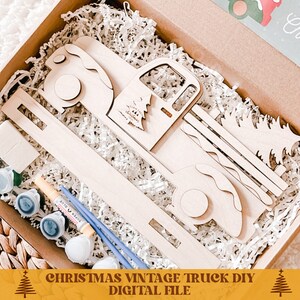 May include: A wooden Christmas vintage truck DIY kit with paint and brushes. The truck is carrying a Christmas tree. The kit is in a brown box with white shredded paper.