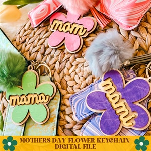 May include: Three flower-shaped keychains with the word "mom" or "mama" in gold letters. The keychains are made of wood and have a light pink, green, and purple color scheme. The keychains are on a woven wicker surface.