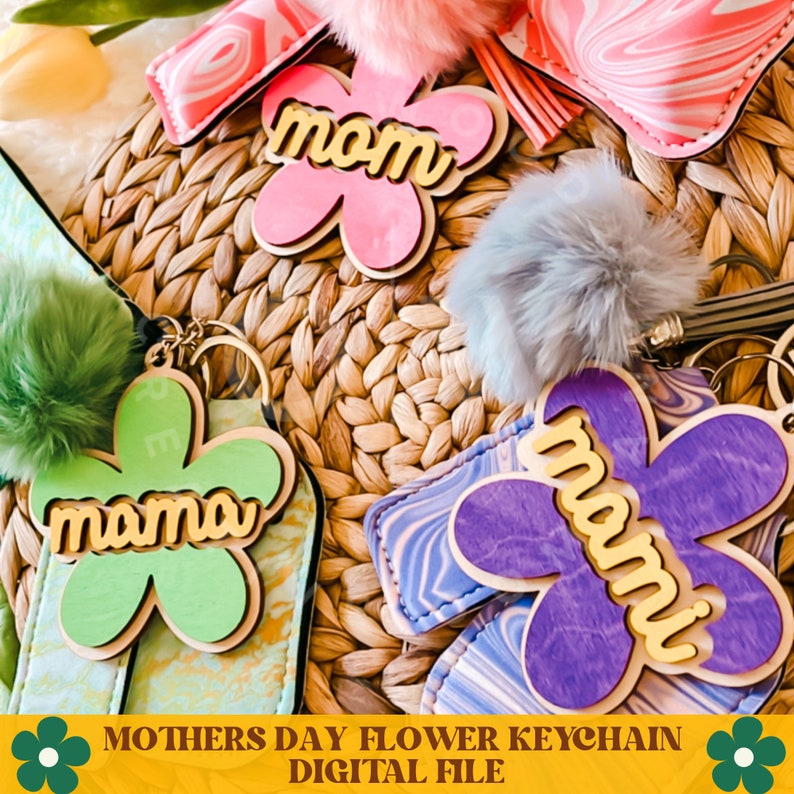 May include: Three flower-shaped keychains with the word "mom" or "mama" in gold letters. The keychains are made of wood and have a light pink, green, and purple color scheme. The keychains are on a woven wicker surface.