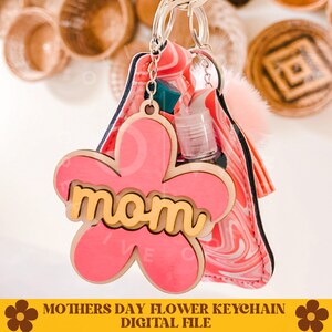 May include: Pink flower-shaped keychain with the word "mom" in yellow letters. The keychain is attached to a pink and white marbled fabric pouch with a clear plastic bottle and a fuzzy white pom-pom.