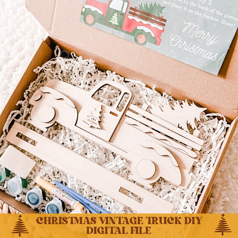 May include: A wooden Christmas truck DIY kit with paint pots and brushes. The truck is carrying a Christmas tree. The kit comes in a brown box with white shredded paper. The box also includes a card with the text "Merry Christmas!" and instructions for assembling the truck.