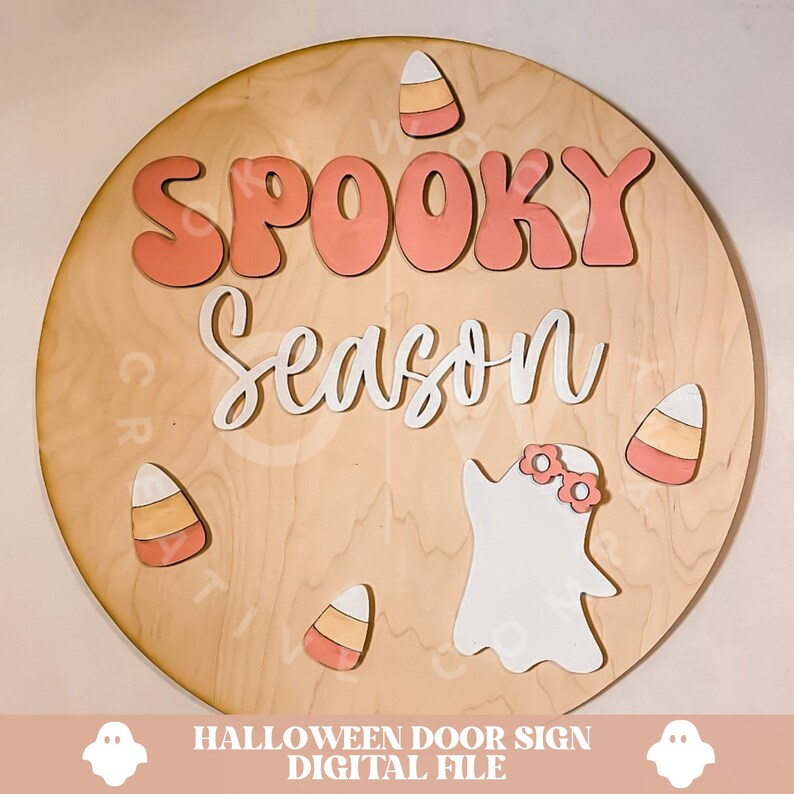 Peut inclure: Une pancarte ronde en bois avec les mots "Spooky Season" en lettres roses et blanches. Il y a quatre d&eacute;coupes de ma&iuml;s sucr&eacute; et une d&eacute;coupe de fant&ocirc;me avec des lunettes roses.