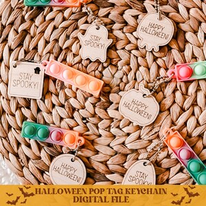 Op de afbeelding: Een set van vijf Halloween-thema pop-it sleutelhangers. De tags zijn gemaakt van hout en hebben verschillende ontwerpen, waaronder een spook, een pompoen en een vleermuis. De tekst op de tags luidt "Stay Spooky!" en "Happy Halloween!"