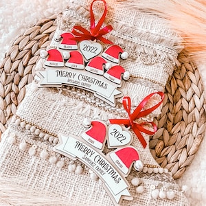 Puede incluir: Dos adornos navide&ntilde;os de madera con gorros de Pap&aacute; Noel rojos y blancos y el texto 'Feliz Navidad' con el nombre de la familia debajo. El adorno superior dice 'Rosado Family' y el inferior 'Sanchez Family'. Ambos adornos tienen el a&ntilde;o '2022' en una pieza con forma de coraz&oacute;n.