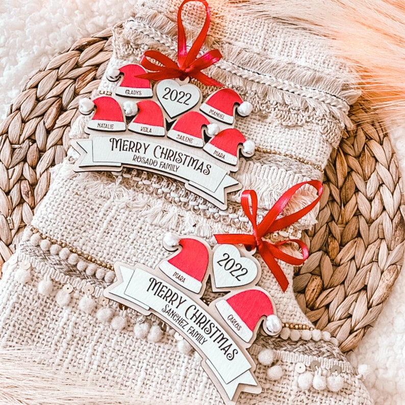 Puede incluir: Dos adornos navide&ntilde;os de madera con gorros de Pap&aacute; Noel rojos y blancos y el texto 'Feliz Navidad' con el nombre de la familia debajo. El adorno superior dice 'Rosado Family' y el inferior 'Sanchez Family'. Ambos adornos tienen el a&ntilde;o '2022' en una pieza con forma de coraz&oacute;n.