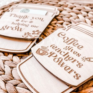 May include: Two wooden coffee cup-shaped ornaments with laser-engraved messages. The first ornament says "Thank you for helping me grow" with a small flower design. The second ornament says "Coffee gives teachers powers" with a small coffee cup design.