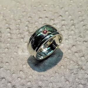 Peut inclure: Une bague en argent avec une bande et une pierre rouge sertie en forme d'étoile. La bague a une surface texturée et une finition polie.