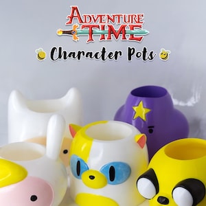 Pode incluir: Uma coleção de vasos de personagens em várias formas e cores, inspirada na série animada Adventure Time. Os vasos apresentam desenhos de Finn, Jake e outros personagens. A imagem inclui o texto "Adventure Time Character Pots."