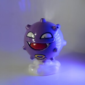 May include: A purple, spherical figurine with a smiling face, white eyes, and a red mouth. The figurine has small spikes and sits atop a white cloud-shaped base. A yellow and black symbol is visible on the base.