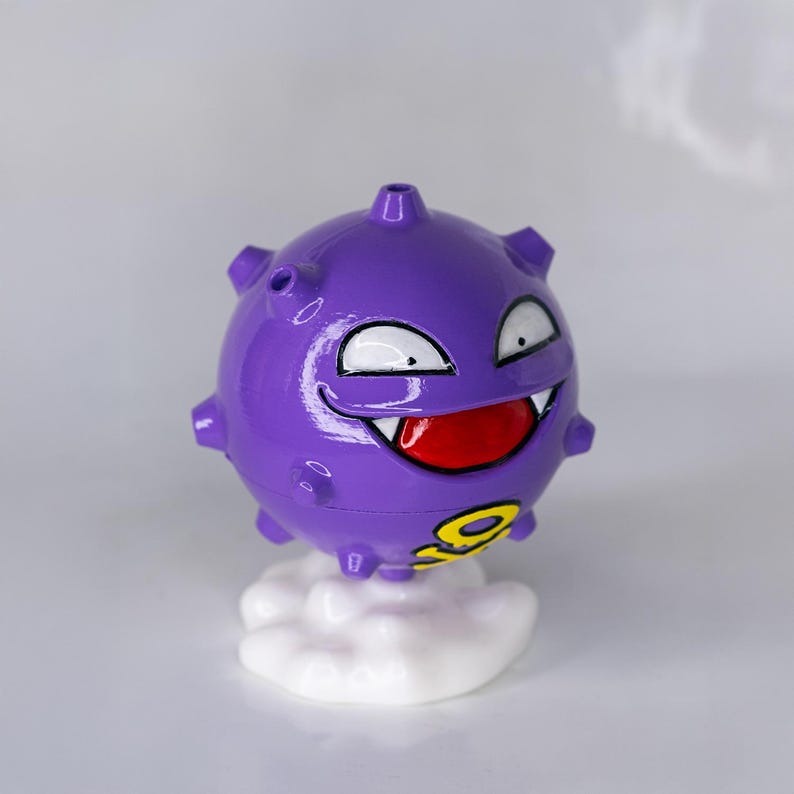 May include: A purple, spherical figurine with a smiling face, white eyes, and a red mouth. The figurine has yellow markings and is mounted on a white cloud-shaped base. The design is reminiscent of a cartoon character.