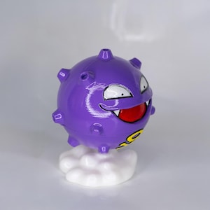 May include: A purple cartoon character figurine with a round body, white eyes, and a red mouth. The figurine is mounted on a white cloud-shaped base. The character has yellow accents.