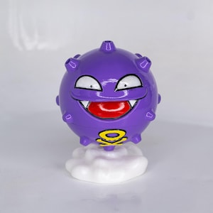 May include: A purple, spherical figurine with a smiling face, white fangs, and a red tongue. The figurine has a yellow and black symbol on its base and sits atop a white cloud-shaped base. The figurine is a cartoon character.