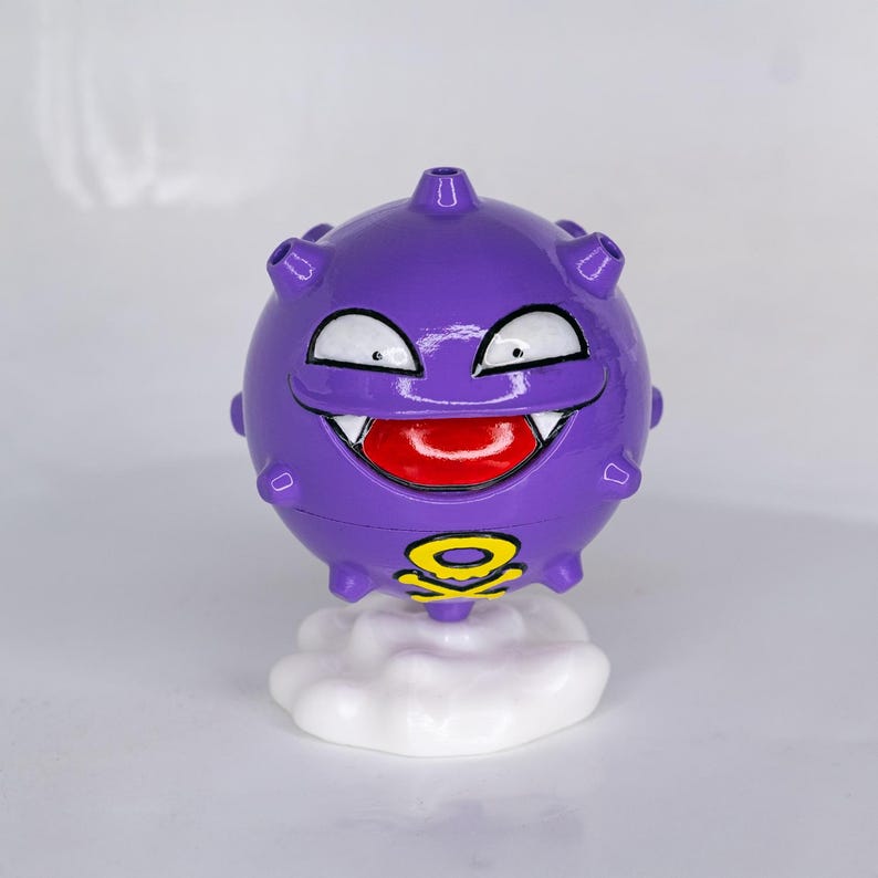 May include: A purple, spherical figurine with a smiling face, white fangs, and a red tongue. The figurine has a yellow and black symbol on its base and sits atop a white cloud-shaped base. The figurine is a cartoon character.