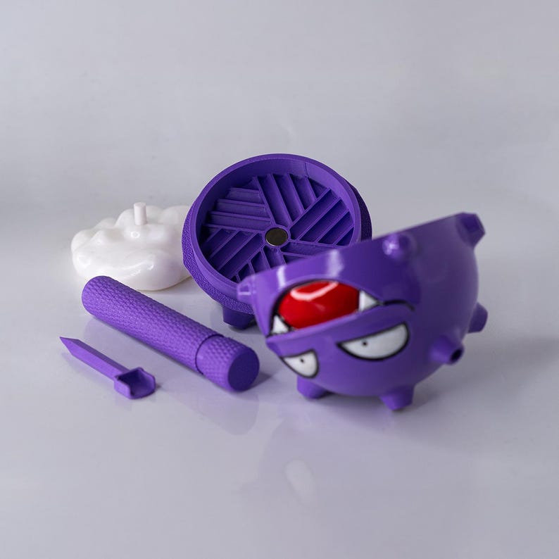 May include: A purple novelty item with a cartoon character design. The item is disassembled, showing a cylindrical piece, a small scoop, a circular piece, and a character-shaped container with a red ball inside. A white, cloud-shaped piece is also present.