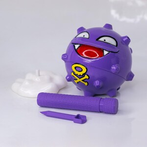 May include: A purple, spherical toy with a smiling face, white teeth, and a red mouth. The toy has a yellow skull and crossbones symbol. A purple cylinder and a purple pointed object are in front of the toy. A white, cloud-shaped stand is behind the toy.