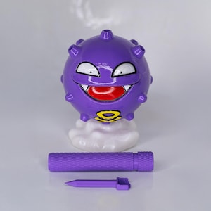 May include: A purple, spherical figurine with a smiling face, white teeth, and a red tongue. It sits atop a white cloud base, with a matching purple cylindrical piece and a pointed tool below. The figurine has a yellow and black symbol.